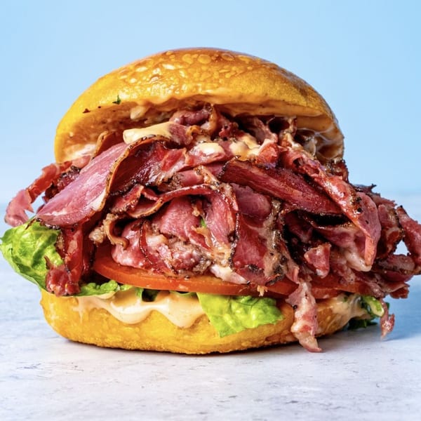 A large pastrami sandwich on a bagel with lettuce, tomato, and sauce, against a blue background.