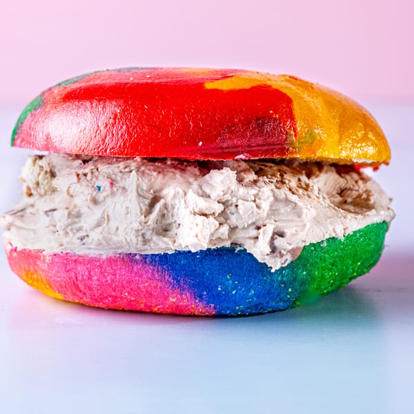 A vibrant rainbow bagel from Beverly Hills Bagel, filled with a generous layer of creamy, light brown spread.