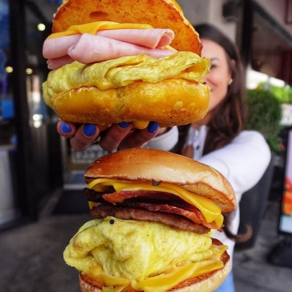 A person holds two large Beverly Hills Bagel breakfast sandwiches with eggs, cheese, and meat.