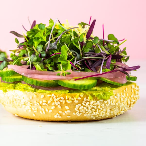 Sesame bagel with avocado spread, cucumber, pickled red onion, and microgreens on a pink background.