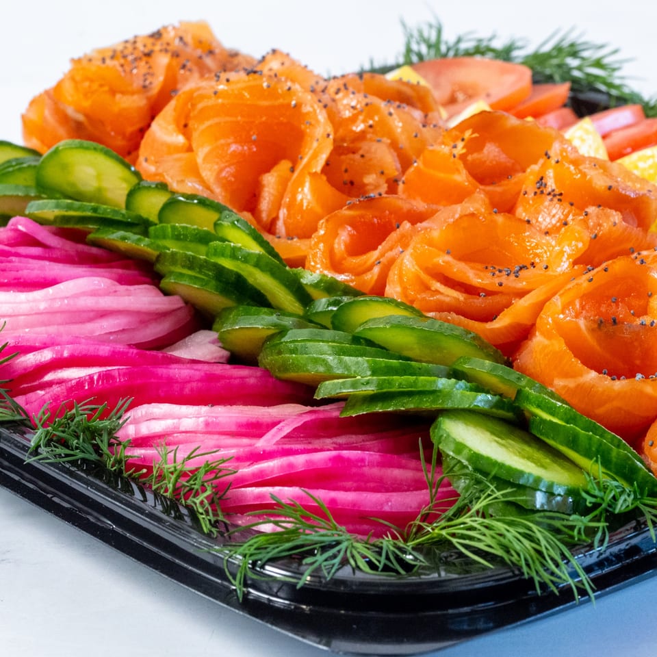 Smoked salmon, sliced cucumbers, pickled red onions, tomatoes, and dill on a black platter.